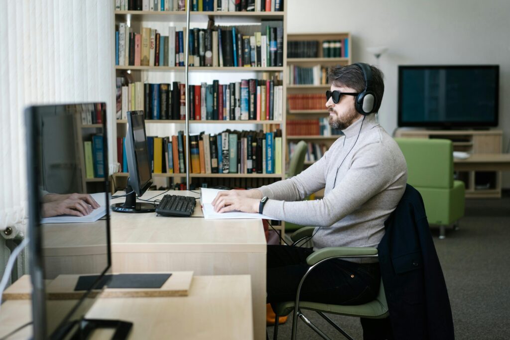 pexels-photo-6981085-6981085 A blind man uses a computer with headphones in a library, highlighting technology's role in accessibility.